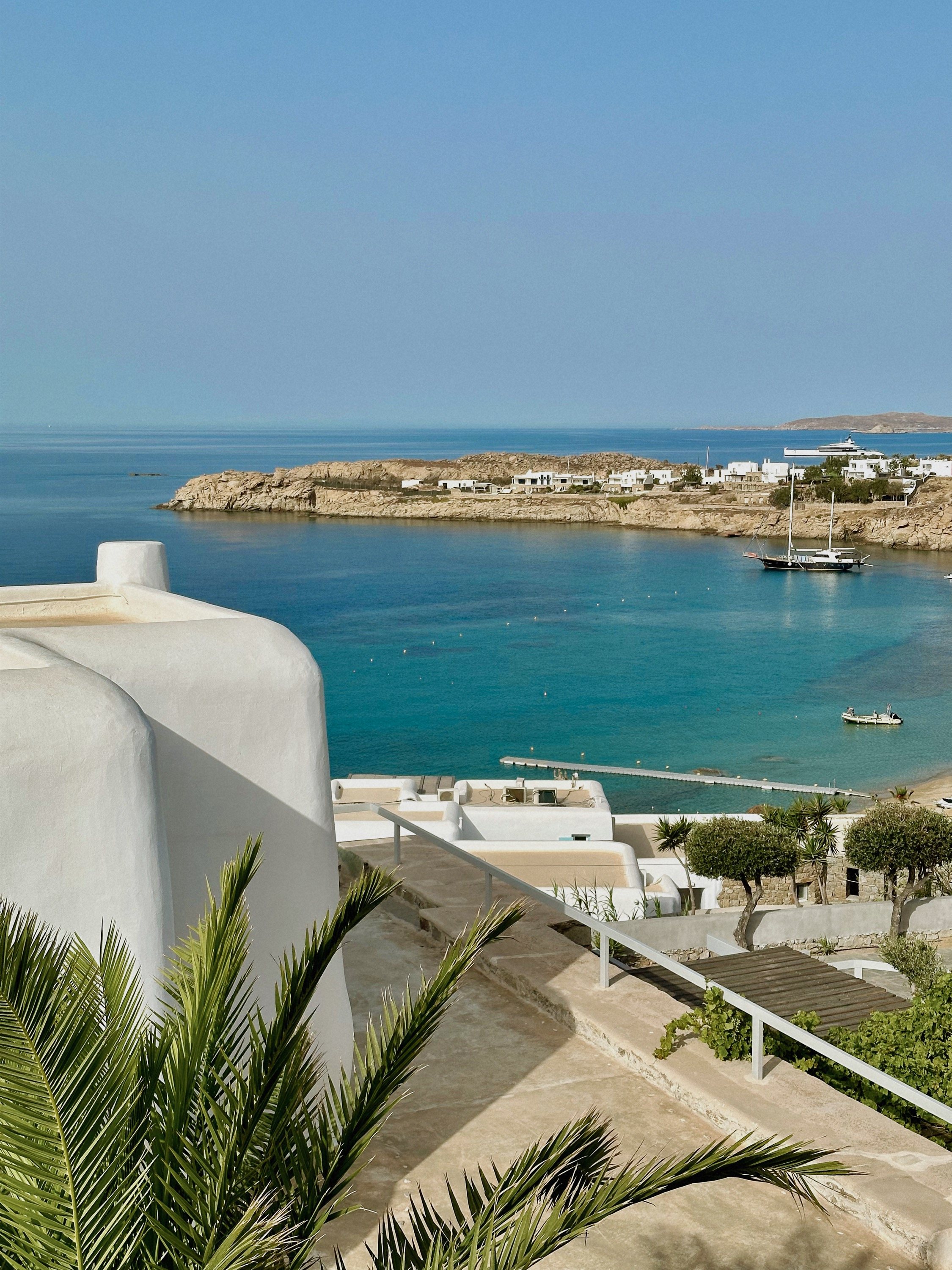 A white Cycladic house and palms above a turquoise bay with sailboats