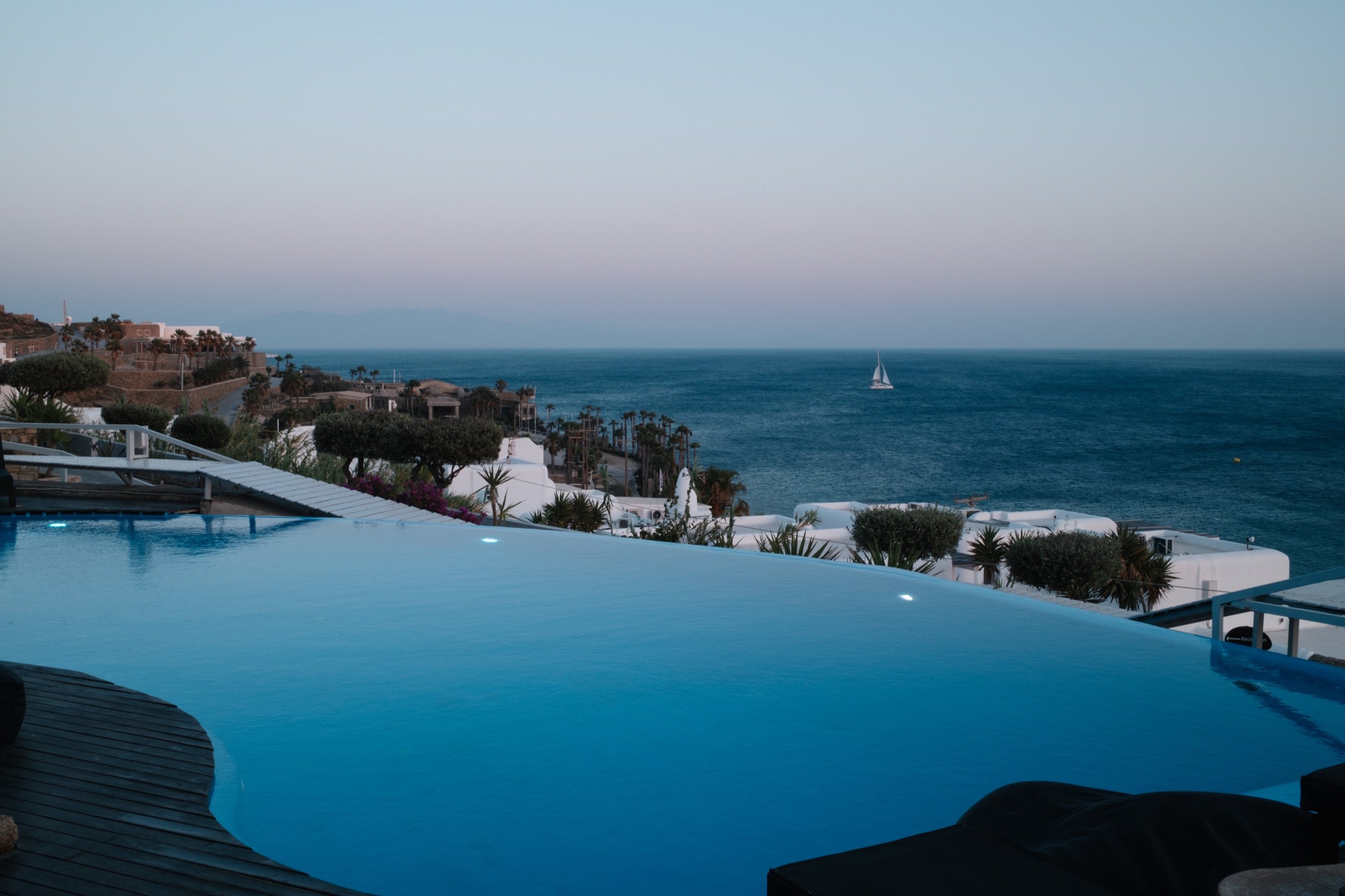An infinity pool at dusk, looking out over the Aegean with a sailboat on the horizon