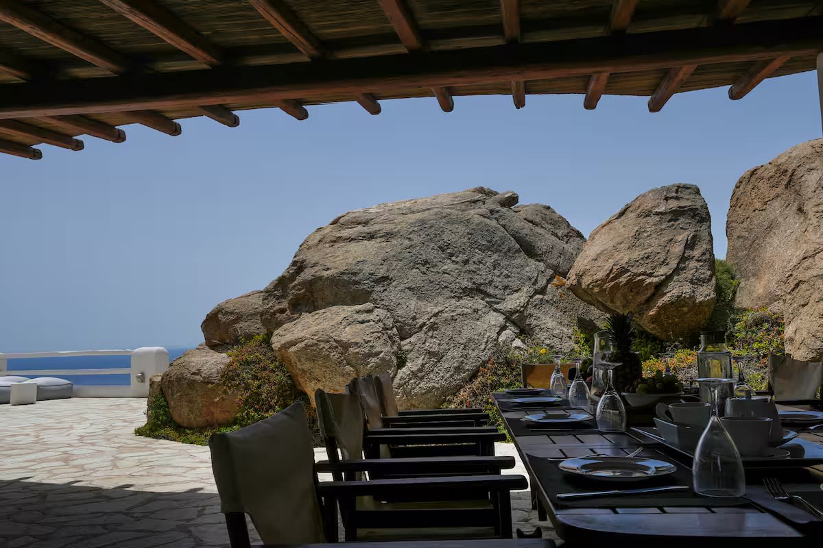 A long dining table set under the pergola, beside the boulders, with the sea beyond